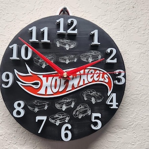 May include: Black round clock with white numerals and red hands. The clock face features the Hot Wheels logo in red and white, with car illustrations. The clock is mounted on a textured wall.