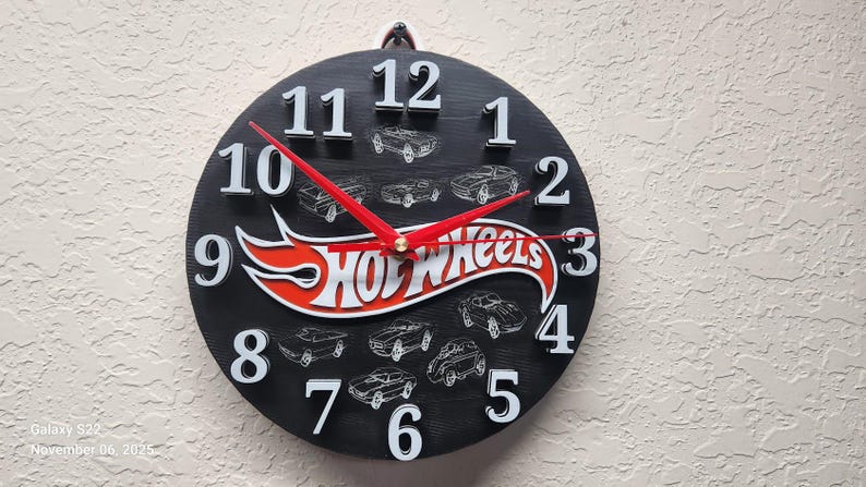 May include: Black round clock with white numerals and red hands. The clock face features the Hot Wheels logo in red and white, with car illustrations. The clock is mounted on a textured wall.