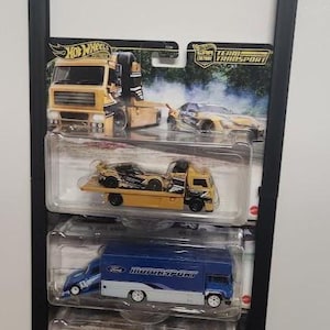 May include: A black wall-mounted display rack holding three Hot Wheels Team Transport die-cast vehicle sets in their original blister packaging. The sets include a yellow race truck, a blue Ford Motorsport hauler, and a white vintage truck with a car.