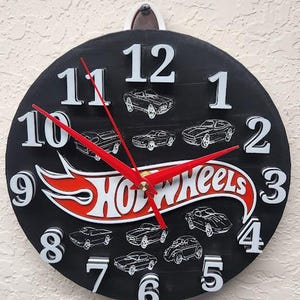 May include: A black round clock with white numbers and red hands. The clock features the Hot Wheels logo in red and white, and illustrations of toy cars. The clock is hanging on a textured wall.
