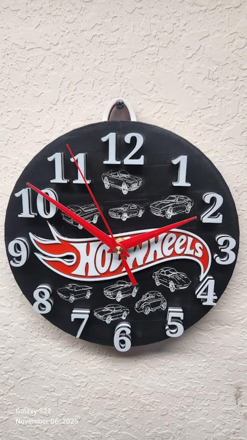 May include: A black round clock with white numbers and red hands. The clock features the Hot Wheels logo in red and white, and illustrations of toy cars. The clock is hanging on a textured wall.