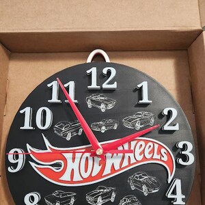May include: Black round clock with white numbers and red hands. The clock features the Hot Wheels logo and car illustrations. The clock is in a cardboard box.