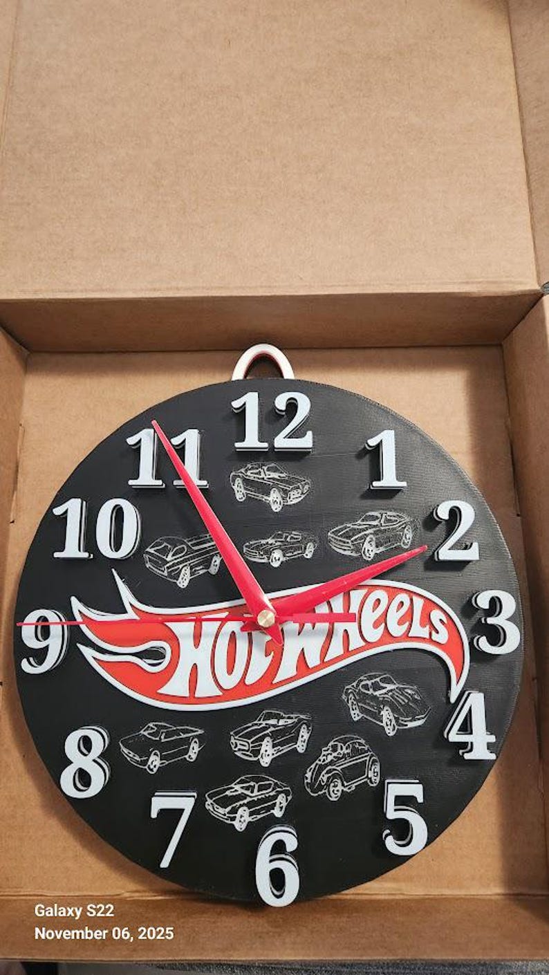 May include: Black round clock with white numbers and red hands. The clock features the Hot Wheels logo and car illustrations. The clock is in a cardboard box.