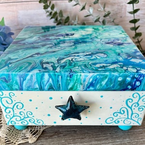May include: A decorative wooden box with a blue and white abstract design on the lid. The box has a white base with teal accents, a star-shaped handle, and teal feet. The box is approximately 6 inches long, 6 inches wide, and 4 inches tall.