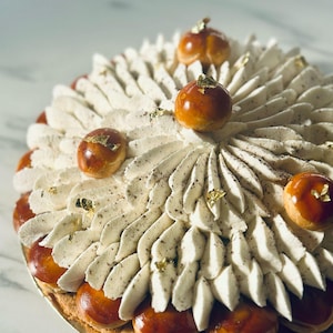 Puede incluir: Una pastelería redonda con glaseado color crema, decorada con esferas glaseadas de caramelo marrón y acentos de pan de oro. La pastelería tiene una base en capas y se presenta en un plato dorado.