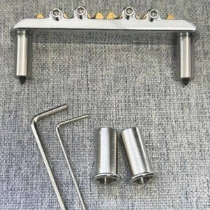 May include: A silver-toned guitar bridge with six adjustable saddles and two posts. Included are two Allen wrenches and two metal sleeves. The bridge has gold-colored accents and is designed for stringed instruments.