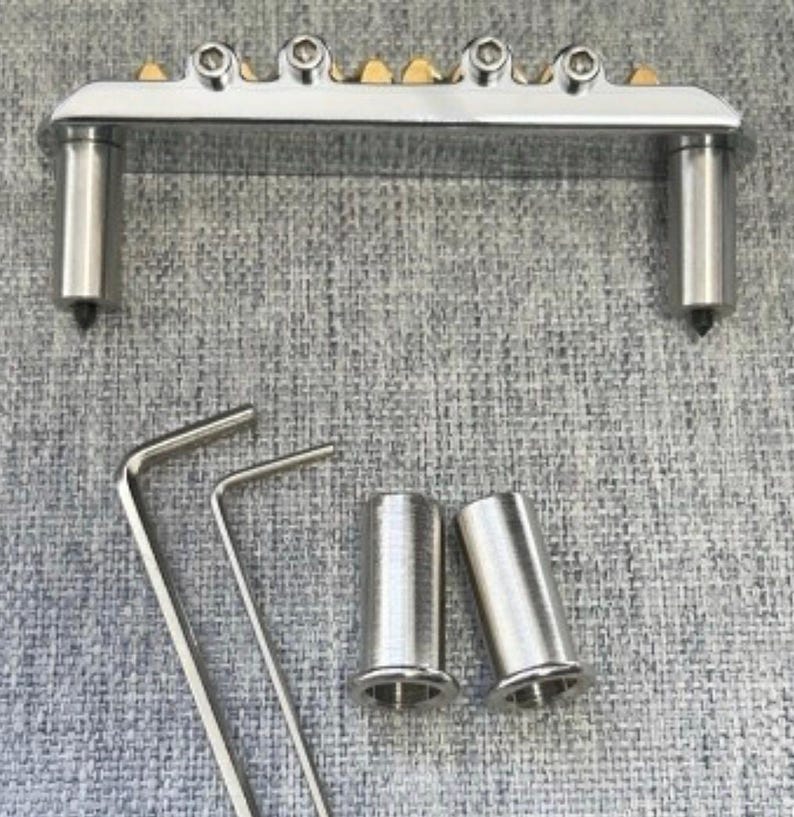 May include: A silver-toned guitar bridge with six adjustable saddles and two posts. Included are two Allen wrenches and two metal sleeves. The bridge has gold-colored accents and is designed for stringed instruments.