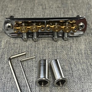 May include: A chrome and gold guitar bridge with adjustable saddles, two Allen wrenches, and two metal bushings. The bridge has a polished finish and is designed for stringed instruments.