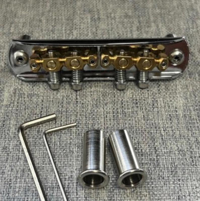 May include: A chrome and gold guitar bridge with adjustable saddles, two Allen wrenches, and two metal bushings. The bridge has a polished finish and is designed for stringed instruments.