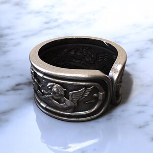 May include: Silver-toned ring with detailed relief carvings. The ring features a design of winged figures and other ornate elements. The interior of the ring is dark, contrasting with the bright exterior. A unique piece of jewelry.