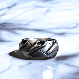 Puede incluir: Un anillo plateado con un diseño tejido. El anillo tiene forma redondeada con una serie de patrones en forma de diamante. Tiene un acabado liso y metálico y está sobre un fondo de mármol.