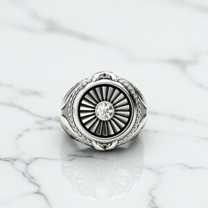 May include: A silver ring with a detailed design featuring a central fan-like pattern and a small, clear gemstone. The ring has an intricate border and a polished finish, set against a marble surface.