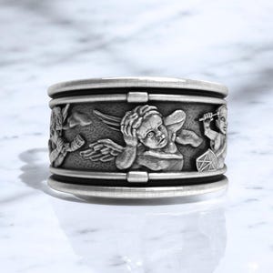 May include: A silver-colored ring with a detailed relief design. The ring features cherubic figures with wings and other decorative elements. The ring has a band-like appearance with a textured surface and a polished finish.