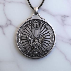 Puede incluir: Collar con colgante plateado con un medallón circular con el texto "ONCE AN EAGLE ALWAYS AN EAGLE". El diseño central incluye un emblema de águila y las letras "BSA". El colgante cuelga de una cadena oscura.