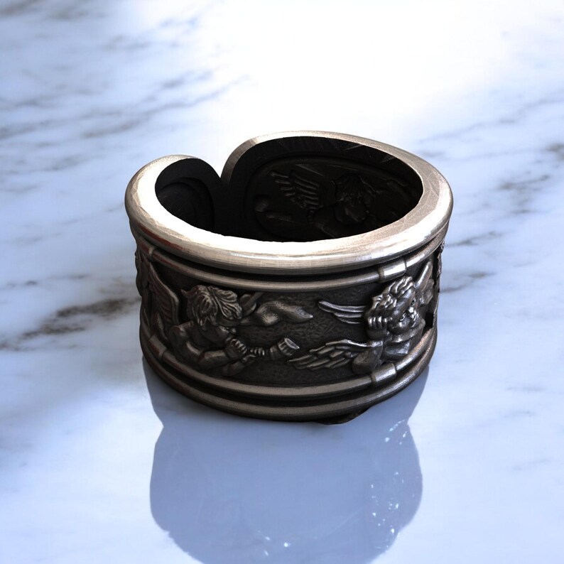May include: A silver-toned ring with an engraved design of cherubs. The ring has a wide band with detailed relief carvings of winged figures. The ring's interior is dark, contrasting with the polished exterior. The ring sits on a white and gray marble surface.