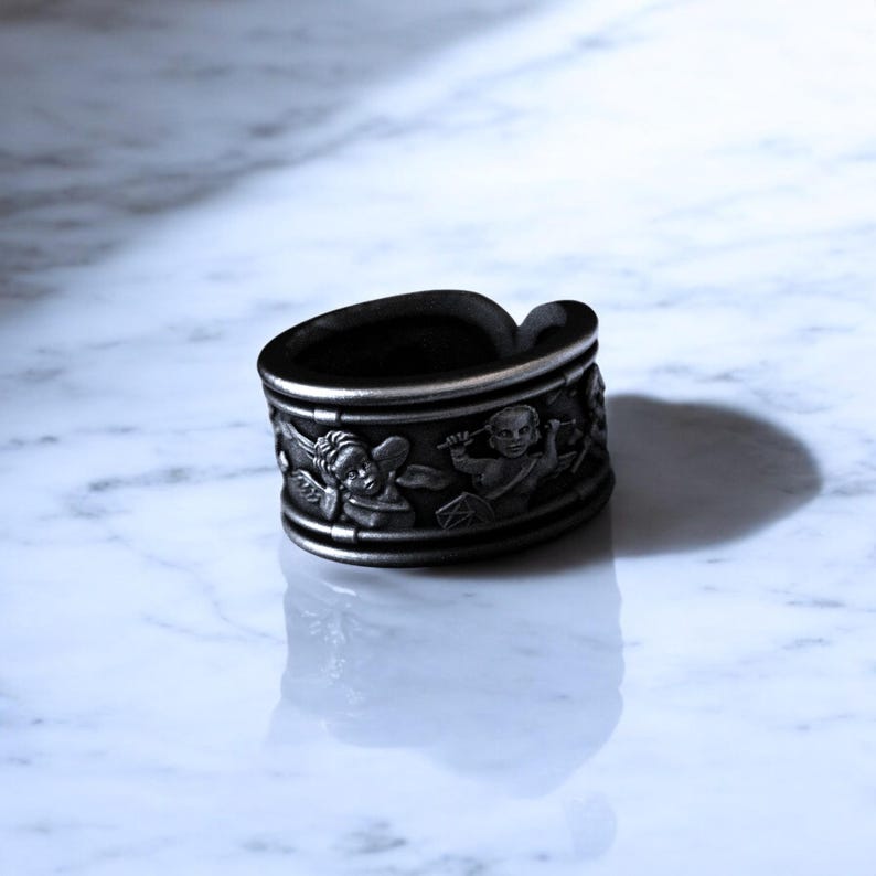May include: A dark gray ring with a detailed relief design. The ring features a band with cherub figures and a slightly curved top. The ring is made of a dark metal and has a matte finish.