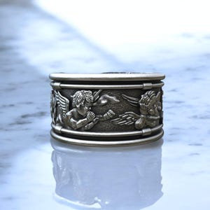 May include: A silver-colored ring with a detailed relief design. The ring features cherubic figures with wings, musical instruments, and other decorative elements. The ring has a wide band and a polished finish.