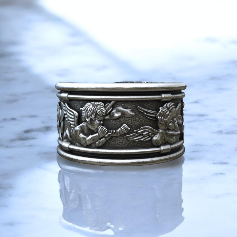 May include: A silver-colored ring with a detailed relief design. The ring features cherubic figures with wings, musical instruments, and other decorative elements. The ring has a wide band and a polished finish.