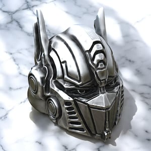 Puede incluir: Una cabeza de robot de color plateado con un diseño detallado. La cabeza tiene un casco grande y angular con una visera y una cara con rasgos afilados.