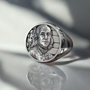 Könnte beinhalten: Ein silberner Ring mit einem detaillierten Relief von Benjamin Franklin. Das Ringdesign umfasst Sterne und Streifen mit poliertem Finish. Der Ring ist ein Statement-Stück, geeignet für den täglichen Gebrauch.