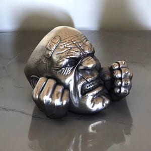 May include: A silver-colored sculpture of a stylized, angry face with clenched fists. The face has a bandage on the forehead and the word "HOOLIGAN" is visible on the teeth. The sculpture is detailed with wrinkles and a textured surface.