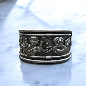 May include: A silver-colored, rectangular box with detailed relief carvings of cherubs. One cherub holds a bow and arrow, while another plays a flute. The box has a hinged lid and a decorative border, showcasing a classical design.