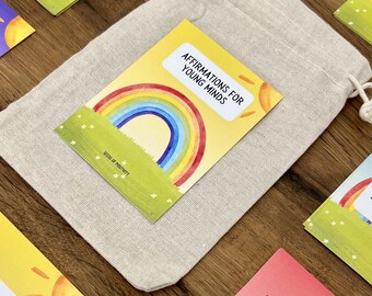 May include: A set of affirmation cards with a rainbow and sun design. The cards are yellow with black text and say "Affirmations for Young Minds" and "Seeds of Positivity". The cards are in a small white drawstring bag.