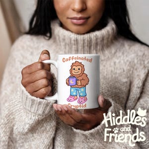 May include: White ceramic mug with a cartoon illustration of a brown Bigfoot wearing blue pajamas and pink slippers. The Bigfoot is holding a purple mug with a crescent moon on it. The text "Caffeinated Cryptid" is printed on the mug.