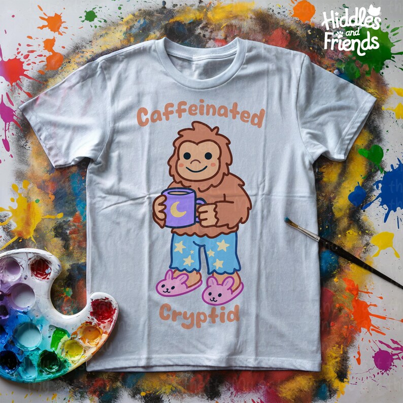 May include: A light blue t-shirt with a cartoon illustration of a brown Bigfoot wearing blue pajamas with stars and holding a purple mug with a crescent moon. The text "Caffeinated Cryptid" is printed on the shirt.