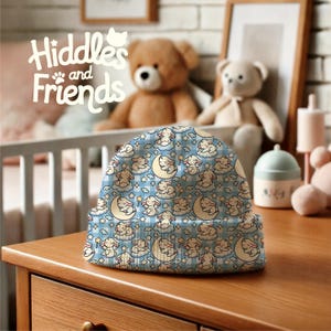 May include: A blue and white beanie with a repeating pattern of sleeping bunnies on clouds and moons. The beanie has a fold-over cuff and is made of soft, comfortable fabric. The text "Hiddles and Friends" is visible in the background.