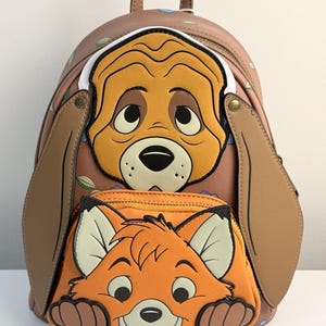 Puede incluir: Una mochila marrón con un diseño de perro y zorro de dibujos animados. La cara del perro está en la parte superior, con orejas grandes y una expresión triste. El zorro está en el bolsillo delantero, con una cara naranja y una expresión sonriente.