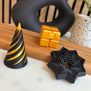May include: Three 3D printed objects: a black and gold cone, a gold cube puzzle, and a black star-shaped object with concentric rings.