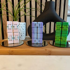 May include: Three fidget toys on a wooden surface. The toys are made of plastic and have a square shape. The first toy is pink and blue, the second is purple and blue, and the third is green. Each toy is on a small silver stand.