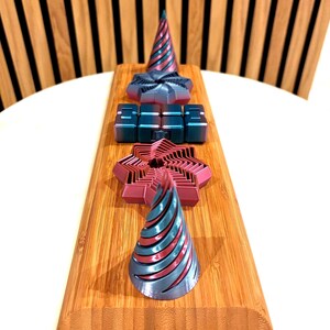 May include: A wooden board showcases various 3D-printed geometric shapes. These include cone-shaped objects with red and silver stripes, star-shaped objects, and cube-like structures in teal and red. The objects are arranged vertically.