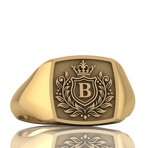 May include: A gold-colored signet ring with a rectangular face. The face features a shield design with a crown, the letter "B", and ornate floral detailing. The ring is highly polished and reflects light.