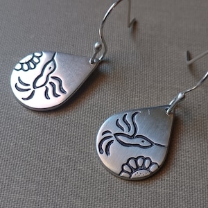 May include: A pair of silver teardrop earrings. Each earring features a hummingbird design with a flower accent. The hummingbird is etched in black, and the earrings have a hook closure.
