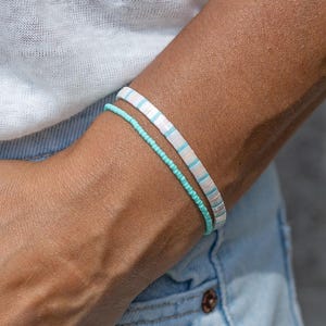 Könnte beinhalten: Zwei Armbänder am Handgelenk. Ein Armband besteht aus kleinen, runden, leuchtend türkisfarbenen Perlen. Das zweite Armband besteht aus flachen, scheibenförmigen Perlen in abwechselnden Farben wie Weiß, Hellblau und Zartrosa für einen Lagenlook.