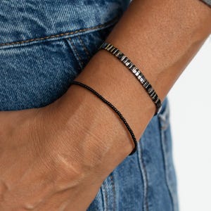 Könnte beinhalten: Zwei Armbänder, die zusammen am Handgelenk getragen werden. Eines ist ein dünnes, zartes schwarzes Perlenarmband, das andere ist ein breiteres, metallisch anthrazitfarbenes Armband aus kleinen, rechteckigen, reflektierenden Gliedern.