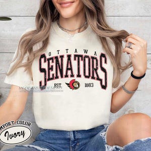 Puede incluir: Una camiseta blanca con un estampado gráfico negro y rojo que dice "Ottawa Senators Est. 1883". La camiseta es de color confort marfil.
