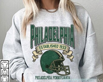 May include: A light grey sweatshirt with a distressed green graphic. The graphic includes the word "PHILADELPHIA", a football helmet, and the text "ESTABLISHED 1933". Below is "PHILADELPHIA, PENNSYLVANIA".