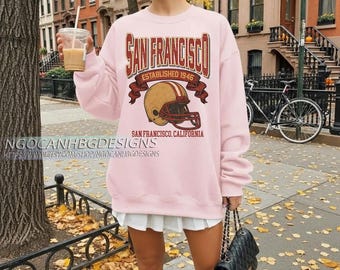 Puede incluir: Sudadera rosa claro con un diseño de San Francisco, que incluye un casco de fútbol y el texto "Established 1946". La sudadera también presenta el texto "San Francisco, California". El nombre y la URL de la tienda Etsy son visibles en la parte inferior.