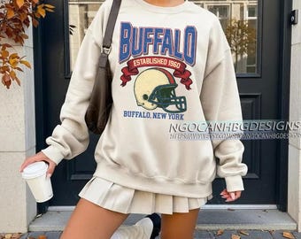 May include: A cream-coloured sweatshirt featuring the word "BUFFALO" in blue, with "ESTABLISHED 1960" and a football helmet graphic. The text "BUFFALO, NEW YORK" is also visible. The sweatshirt has long sleeves.