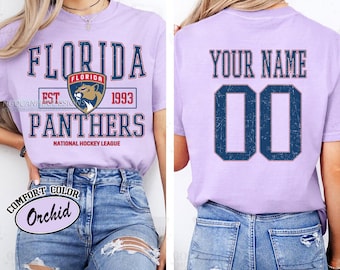 May include: A light purple t-shirt with the text "FLORIDA PANTHERS" in navy blue, along with a team logo and the text "NATIONAL HOCKEY LEAGUE." The back of the shirt has "YOUR NAME" and the number "00" in navy blue.