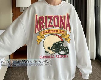 Op de afbeelding: Witte sweatshirt met een grafisch ontwerp met het woord "ARIZONA" in bordeauxrood en goud, een footballhelm en de tekst "ESTABLISHED 1920" en "GLENDALE, ARIZONA". De sweatshirt heeft ook de tekst "NGOCANHBGDESSIGNS" en een websiteadres.
