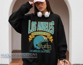 May include: Black sweatshirt featuring a teal and gold graphic that reads "Los Angeles Established 1960" above a teal football helmet. The text "Los Angeles, California" is below the helmet. The shop name is at the bottom.