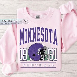 May include: A pale pink sweatshirt featuring the word "MINNESOTA" in purple block letters. Below the word is a purple football helmet, the numbers "1961", and the word "FOOTBALL". The sweatshirt has long sleeves.