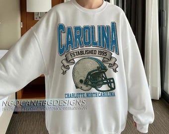 Może przedstawiać: Biała bluza z niebiesko-turkusową grafiką z napisem "CAROLINA" nad kaskiem do futbolu amerykańskiego, tekstem "ESTABLISHED 1995" i "CHARLOTTE, NORTH CAROLINA". Bluza ma również napis "NGOCANHBGD ESIGNS" na dole.