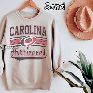 May include: A light gray sweatshirt with the text "Carolina Hurricanes" in black and red lettering. The text is surrounded by a black and white stripe design. The sweatshirt has a crew neck and long sleeves.