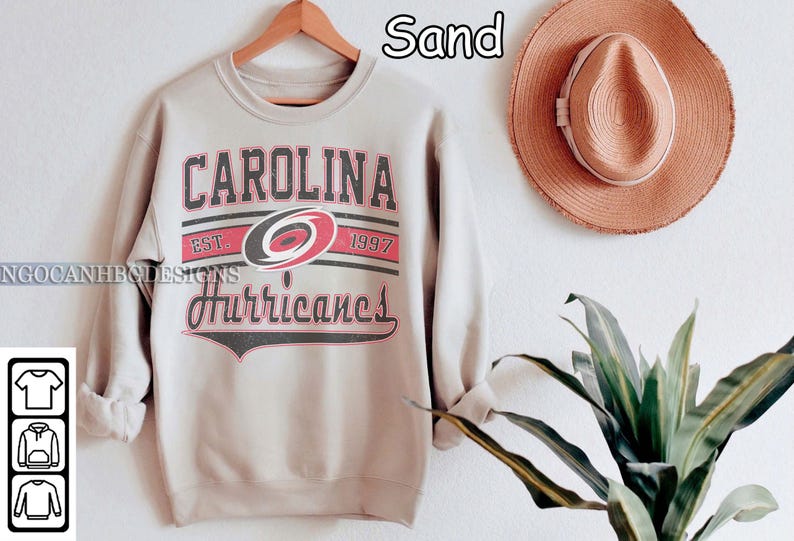 May include: A light gray sweatshirt with the text "Carolina Hurricanes" in black and red lettering. The text is surrounded by a black and white stripe design. The sweatshirt has a crew neck and long sleeves.