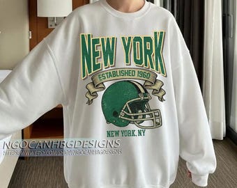 Könnte beinhalten: Weißes Sweatshirt mit einem grün-goldenen "NEW YORK"-Aufdruck, "ESTABLISHED 1960" und einem grünen Footballhelm. Das Design enthält auch "NEW YORK, NY". Ein lässiges, bequemes Kleidungsstück.